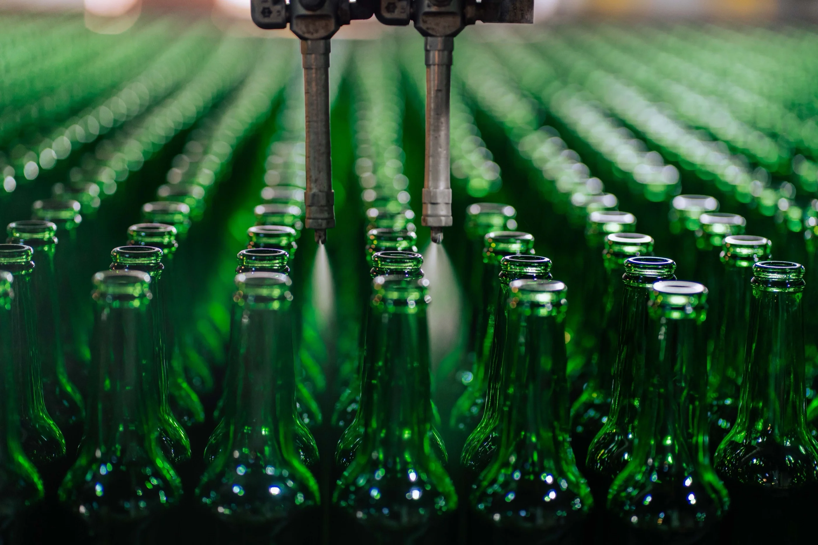 Rows of empty green glass bottles on a conveyor pass under spray nozzles