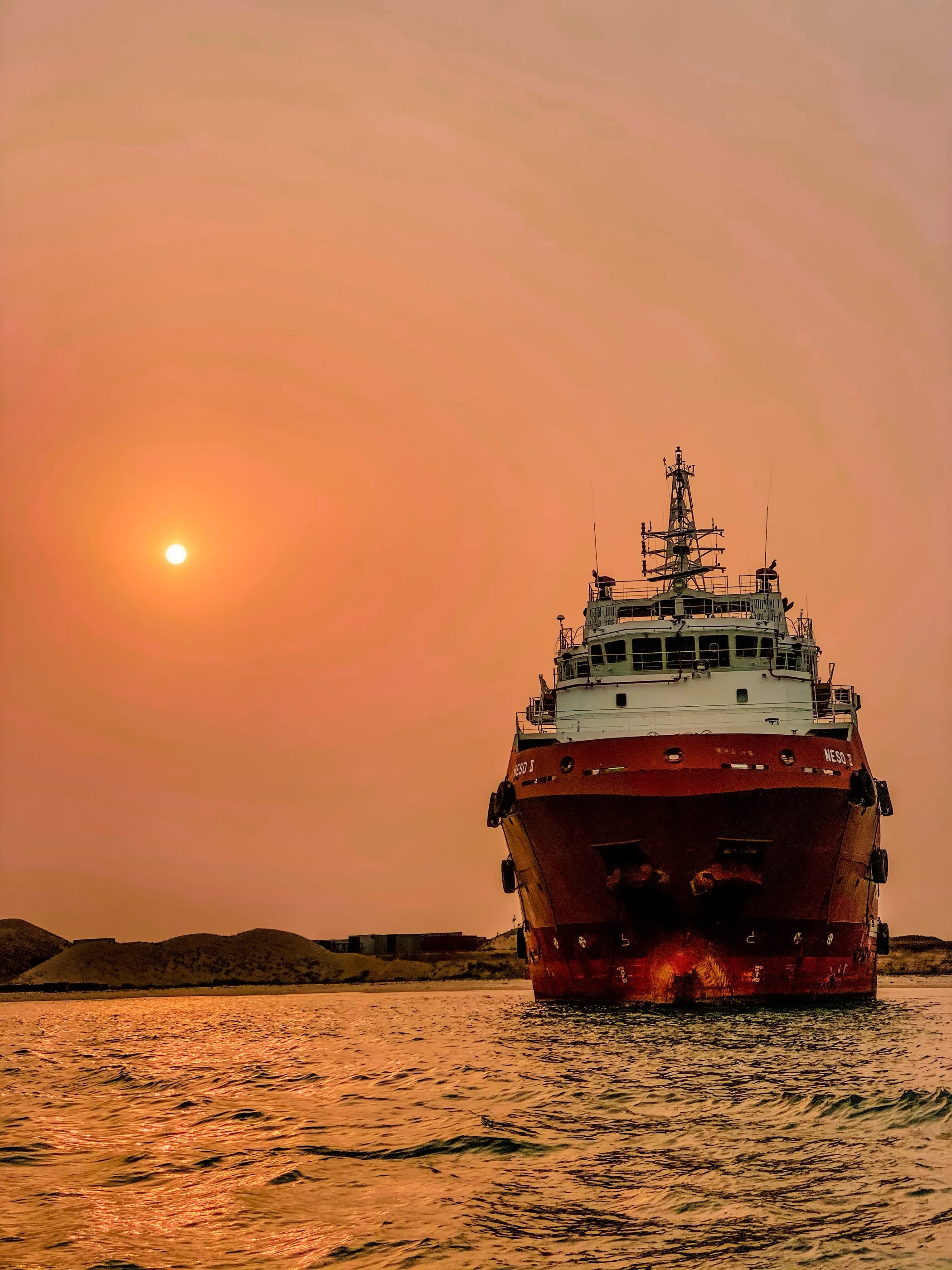A red-hulled offshore supply vessel in Lagos harbour under a hazy orange sunset