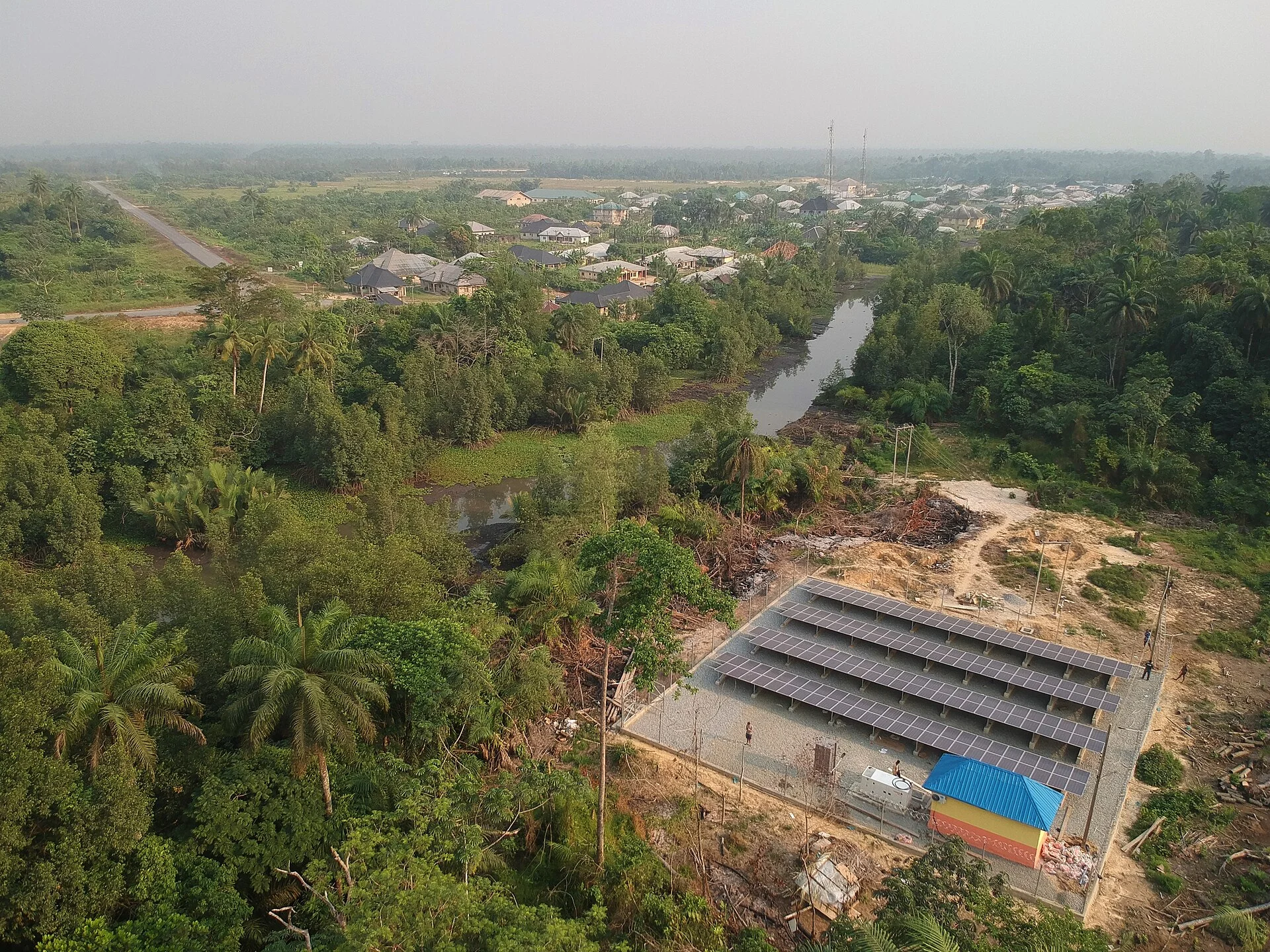 Aerial view of a fenced solar mini-grid array cut into dense palm forest beside a creek in Bayelsa