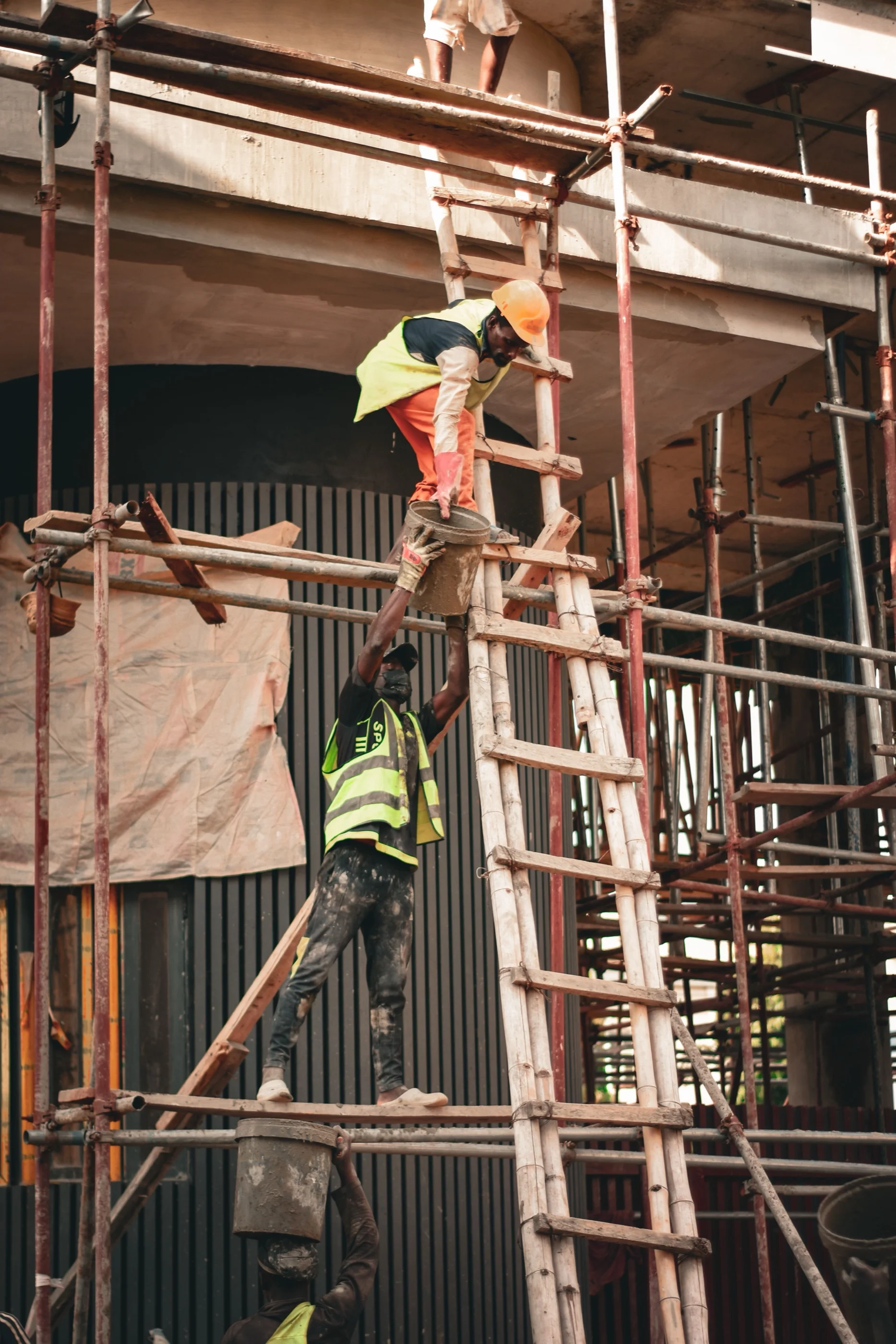 Construction workers in hard hats and hi-vis vests pass a bucket up a ladder on scaffolding in Nigeria