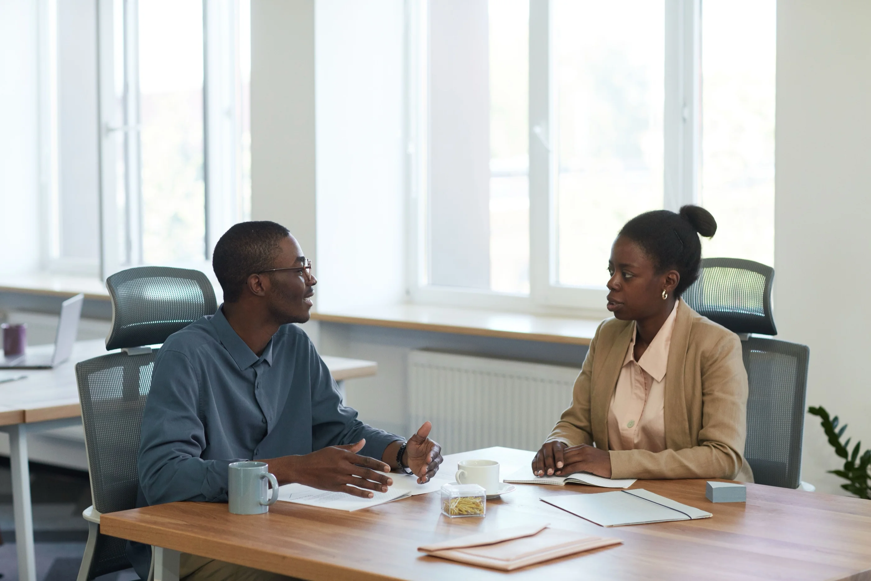 A man and a woman talk across a wooden desk over open notebooks in a bright office