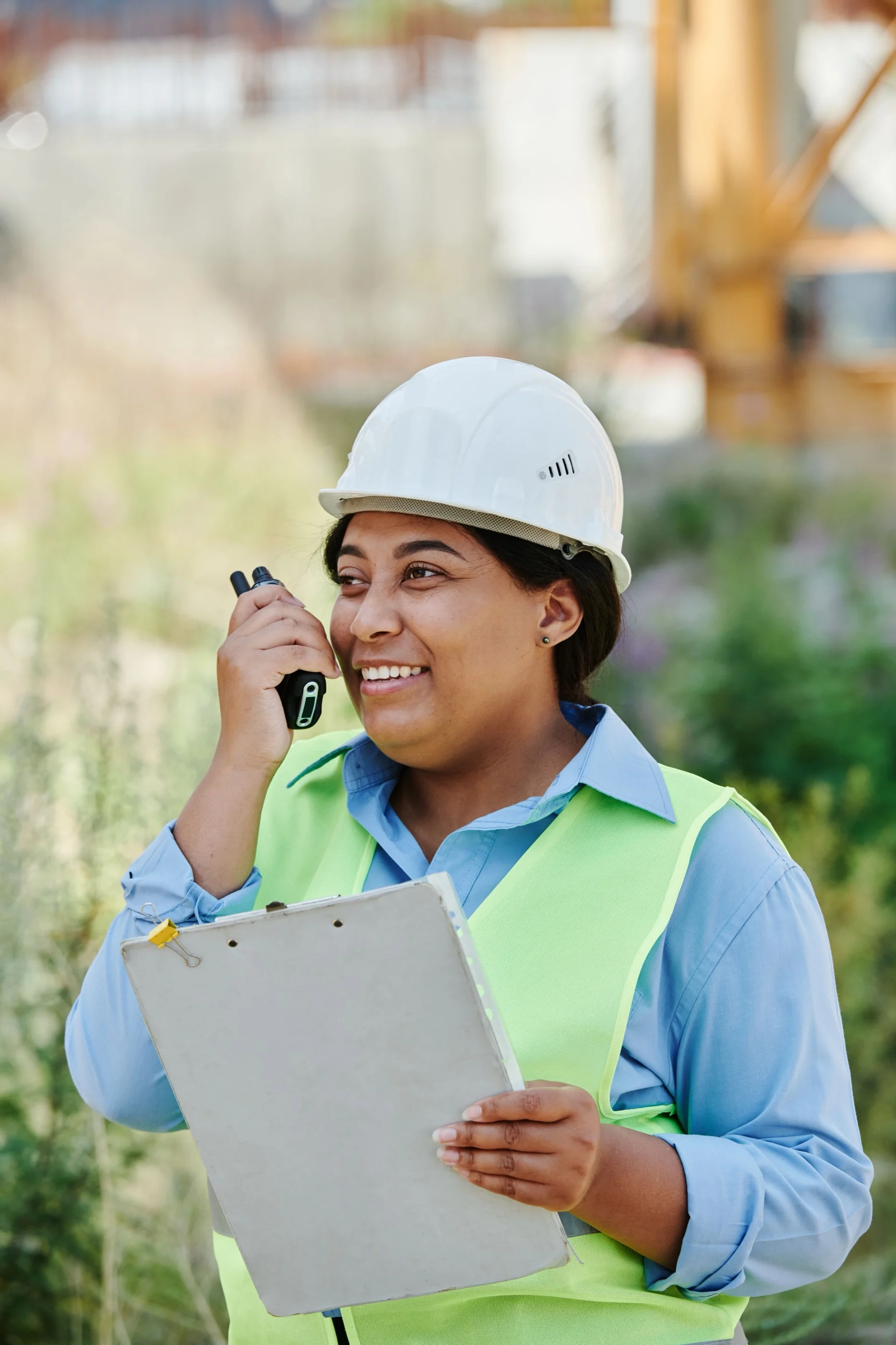 A woman in a white hard hat and hi-vis vest holds a clipboard and speaks into a radio outdoors